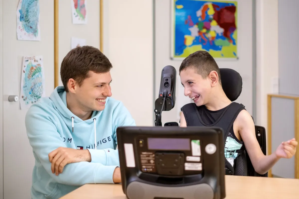 Ein Schulbegleiter hilft einem Sch&uuml;ler im Rollstuhl, sie lachen zusammen