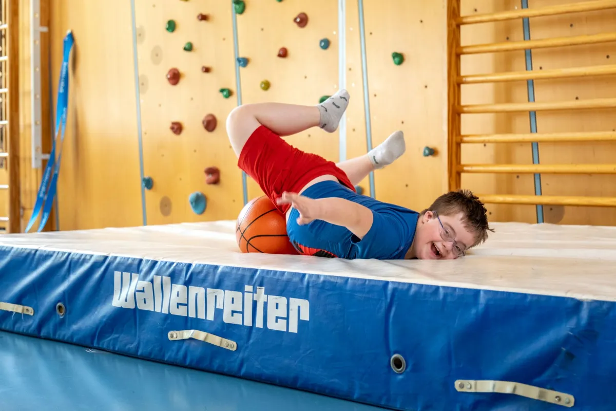 Schüler turnt auf einer dicken Sportmatte in der Turnhalle und spielt mit einem Basketball