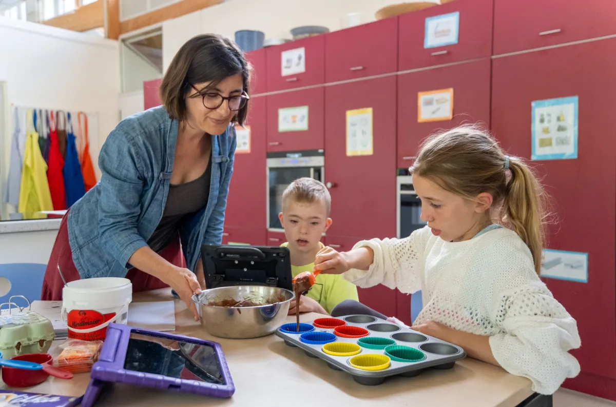 Zwei Schulkinder backen mit ihrer Lehrerin Muffins, sie füllen den Teig in die Förmchen